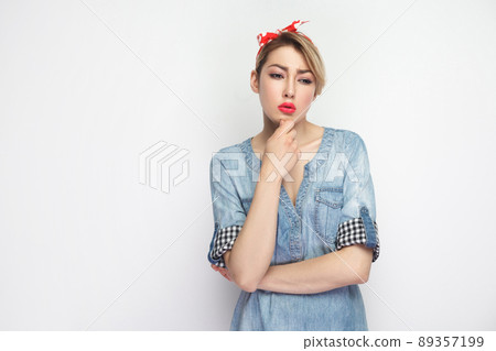 Portrait of thoughtful beautiful young woman in casual blue denim shirt with makeup and red headband standing touching her chin, thinking what to do. indoor studio shot, isolated on white background. Portrait of thoughtful beautiful young woman in casual blue denim shirt with makeup and red headband standing touching her chin, thinking what to do. indoor studio shot, isolated on white background. 89357199