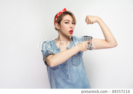 Portrait of strong proud beautiful young woman in casual blue denim shirt with makeup, red headband standing looking at camera and pointing at her biceps hand. studio shot isolated on white background 89357218
