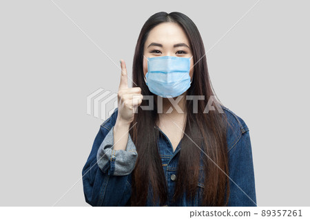 Portrait of exicted beautiful brunette asian girl with surgical medical mask in blue denim jacket standing and looking at camera , finger up. indoor studio shot, isolated on grey background. Portrait of exicted beautiful brunette asian girl with surgical medical mask in blue denim jacket standing and looking at camera , finger up. indoor studio shot, isolated on grey background. 89357261