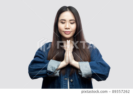 Portrait of calm beautiful brunette asian young woman in casual blue denim jacket with makeup standing with palm hands and yoga meditating. indoor studio shot, isolated on light grey background. 89357262