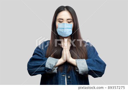 Portrait of calm beautiful brunette asian young woman with surgical medical mask in blue denim jacket standing with palm hands and yoga meditating. indoor studio shot, isolated on grey background. 89357275
