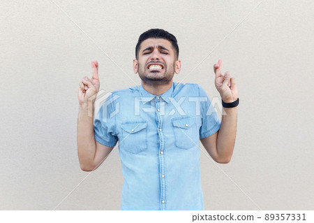 Portrait of hopeful handsome young bearded man in blue shirt standing with crossed fingers, clenching teeth and hope to win. indoor studio shot isolated on light beige wall background. 89357331