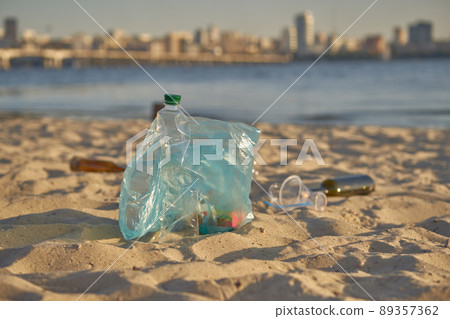 Blue bag with garbage lies on beach of picturesque river, crumpled plastic and glass bottles are scattered nearby. Cleaning up trash. Close-up. Blue bag with garbage lies on beach of picturesque river, crumpled plastic and glass bottles are scattered nearby. Cleaning up trash. Close-up. 89357362