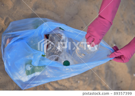 Young volunteer in purple gloves is walking with garbage bag along a dirty beach of the river and cleaning up trash. People and ecology. Close-up. 89357399