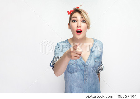 Wow, its unbelievable. Portrait of shocked beautiful young woman in casual blue shirt with makeup and red headband standing and pointing at camera. indoor studio shot, isolated on white background. Wow, its unbelievable. Portrait of shocked beautiful young woman in casual blue shirt with makeup and red headband standing and pointing at camera. indoor studio shot, isolated on white background. 89357404