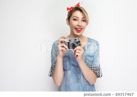 Portrait of beautiful young blogger woman in casual blue denim shirt with makeup and red headband standing, holding retro camera with toothy smile. indoor studio shot, isolated on white background. Portrait of beautiful young blogger woman in casual blue denim shirt with makeup and red headband standing, holding retro camera with toothy smile. indoor studio shot, isolated on white background. 89357407