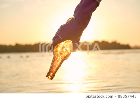 Volunteer hand in purple glove takes out a used glass bottle from the river. Sunset, green trees. People and ecology. Riverside pollution. Close-up. 89357445