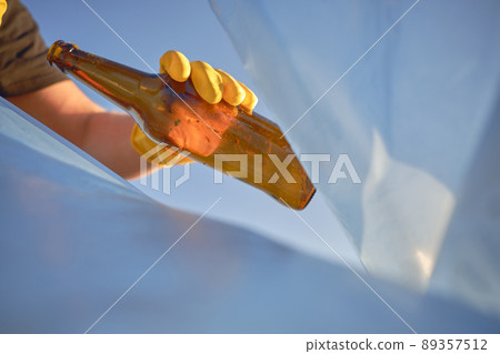 Hand of a conscious person in a yellow rubber glove puts a used brown glass bottle in a blue trash bag. Blue sky. Riverside pollution. Close-up shot. 89357512