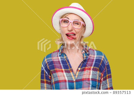 Portrait of funny crazy modern stylish mature woman in casual style with hat and eyeglasses standing, winking, tongue out and looking at camera. indoor studio shot isolated on yellow background. 89357513
