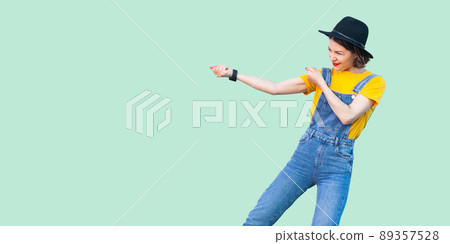 Profile side view portrait of pretty young hipster girl in blue denim overalls, yellow shirt and black hat standing with fist or pulling gesture. indoor studio shot isolated on light green background. Profile side view portrait of pretty young hipster girl in blue denim overalls, yellow shirt and black hat standing with fist or pulling gesture. indoor studio shot isolated on light green background. 89357528