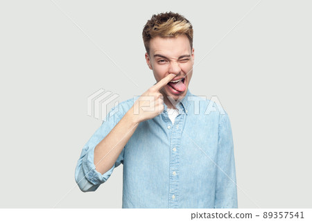 Portrait of crazy handsome young man in light blue shirt standing, drilling his nose, winking, tongue out and looking at camera with funny face. indoor studio shot on grey background copy space. Portrait of crazy handsome young man in light blue shirt standing, drilling his nose, winking, tongue out and looking at camera with funny face. indoor studio shot on grey background copy space. 89357541
