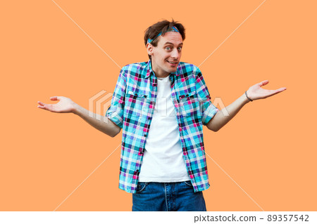I don't know. Portrait of confused young man in casual blue checkered shirt headband standing raised arms and looking at camera with doubtful face. indoor studio shot, isolated on orange background. 89357542
