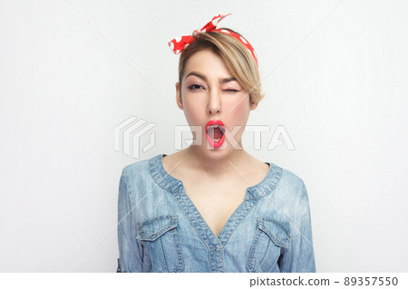 Portrait of funny beautiful young woman in casual blue denim shirt with makeup and red headband standing winking and looking at camera with surprised face. studio shot, isolated on white background. 89357550