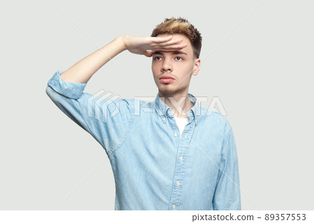 Portrait of serious attentive handsome young man in light blue shirt standing with hand on forehead and looking away with serious face. indoor studio shot on grey background copy space. 89357553