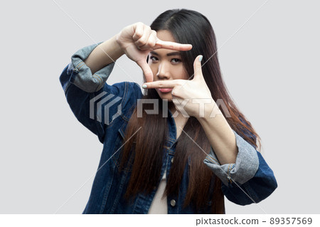 Portrait of serious beautiful brunette asian young woman in blue denim jacket with makeup standing and looking at camera with crop composition gesture. studio shot, isolated on light grey background. Portrait of serious beautiful brunette asian young woman in blue denim jacket with makeup standing and looking at camera with crop composition gesture. studio shot, isolated on light grey background. 89357569