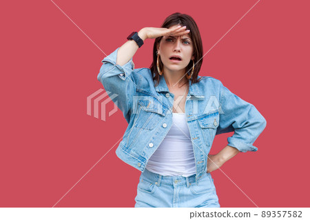 Portrait of attentive beautiful brunette young woman with makeup in denim casual style standing seriously, hand on waist looking with hand on forehead. indoor studio shot, isolated on red background. Portrait of attentive beautiful brunette young woman with makeup in denim casual style standing seriously, hand on waist looking with hand on forehead. indoor studio shot, isolated on red background. 89357582