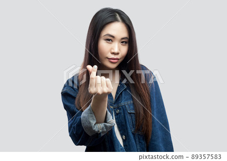 Portrait of funny beautiful brunette asian young woman in casual blue denim jacket with makeup standing and showing money gesture and looking at camera. studio shot, isolated on light grey background. Portrait of funny beautiful brunette asian young woman in casual blue denim jacket with makeup standing and showing money gesture and looking at camera. studio shot, isolated on light grey background. 89357583