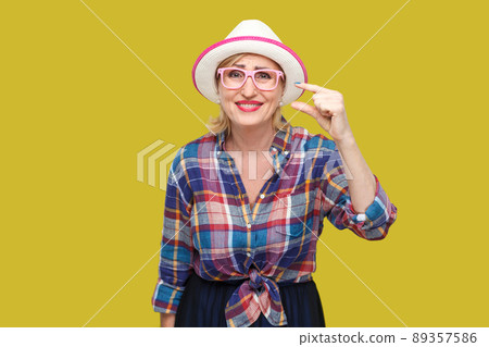 I need a little more. Portrait of pleased modern stylish mature woman in casual style with hat and eyeglasses standing and asking for something more. indoor studio shot isolated on yellow background. 89357586