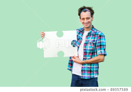 Happy handsome young man in checkered shirt standing and holding large piece of puzzle, looking at camera with toothy smile. Indoor, isolated, copy space, green background, studio shot 89357589