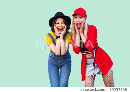 Portrait of two best friend beautiful amazing happy fashionable hipster girls standing and screaming with unbelievable face, looking at camera. Indoor studio shot, isolated on green background. Portrait of two best friend beautiful amazing happy fashionable hipster girls standing and screaming with unbelievable face, looking at camera. Indoor studio shot, isolated on green background. 89357596
