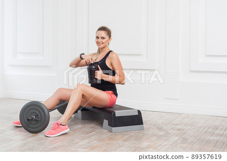 Happy beautiful young athletic causcasian woman pink shorts and black top siting, drink water and have a rest after hard working on the gym. Indoor, studio shot, sport concept, looking at camera Happy beautiful young athletic causcasian woman pink shorts and black top siting, drink water and have a rest after hard working on the gym. Indoor, studio shot, sport concept, looking at camera 89357619