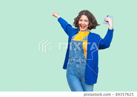Beautiful joyful young adult woman in hipster wear in denim overalls standing, holding phone and listening favourite music with handsets and dancing, looking at camera. Studio shot, green background Beautiful joyful young adult woman in hipster wear in denim overalls standing, holding phone and listening favourite music with handsets and dancing, looking at camera. Studio shot, green background 89357625