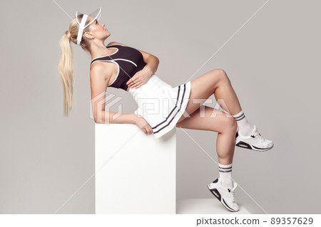 Side view portrait of sporty young athletic woman in fashionable tennis sportwear siting on white cube and posing beautiful perfect figure. Studio shot, indoor, grey background. Sporty concept. Side view portrait of sporty young athletic woman in fashionable tennis sportwear siting on white cube and posing beautiful perfect figure. Studio shot, indoor, grey background. Sporty concept. 89357629