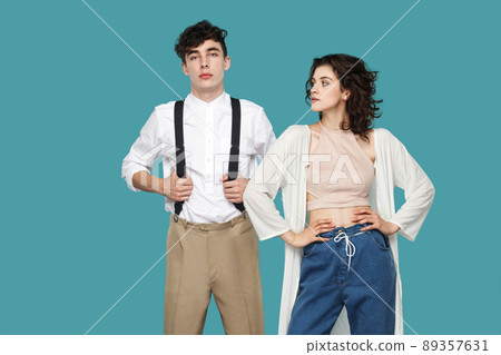 Portrait of two serious proud brunette stylish partner or couple standing, hands on waist, looking with confident. success of partnership or friendship. indoor studio shot isolated on blue background. 89357631