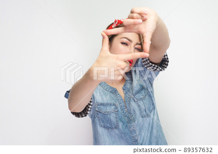 Portrait of focused beautiful young woman in casual blue denim shirt with red headband standing with crop composition gesture and looking at camera. indoor studio shot, isolated on white background. Portrait of focused beautiful young woman in casual blue denim shirt with red headband standing with crop composition gesture and looking at camera. indoor studio shot, isolated on white background. 89357632