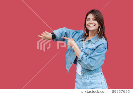 Portrait of happy beautiful brunette young woman with makeup in denim casual style standing showing and pointing at her smart watch with toothy smile. indoor studio shot, isolated on red background. Portrait of happy beautiful brunette young woman with makeup in denim casual style standing showing and pointing at her smart watch with toothy smile. indoor studio shot, isolated on red background. 89357635