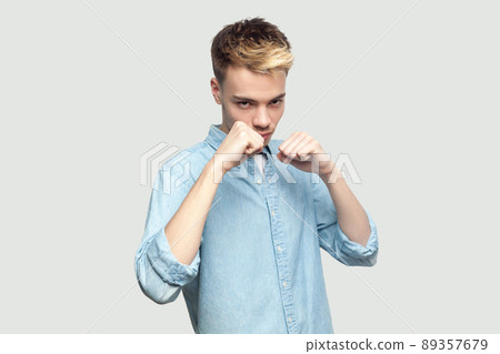 Portrait of serious handsome young man in light blue shirt standing in boxing fists, looking at camera and ready to attack or defence. indoor studio shot on grey background copy space. Portrait of serious handsome young man in light blue shirt standing in boxing fists, looking at camera and ready to attack or defence. indoor studio shot on grey background copy space. 89357679