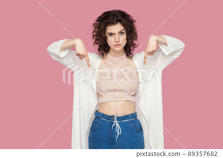 Here and right now. Portrait of serious cute brunette young woman with curly hairstyle in casual style standing and pointing down and looking at camera. indoor studio shot isolated on pink background. Here and right now. Portrait of serious cute brunette young woman with curly hairstyle in casual style standing and pointing down and looking at camera. indoor studio shot isolated on pink background. 89357682