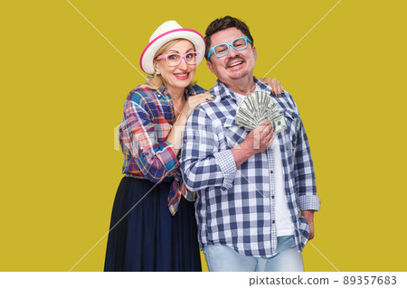 Happy wealthy family, adult man and woman in casual checkered shirt standing pickaback together, holding fan of dollar, toothy smile, looking at camera. Indoor,isolated, studio shot, yellow background 89357683
