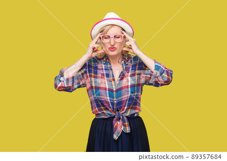 Headache or confusion. Portrait of frowning stylish mature woman in casual style with hat and eyeglasses standing holding her head with head pain. indoor studio shot isolated on yellow background. 89357684