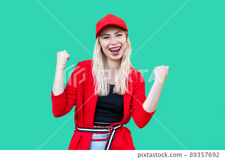 Portrait of happy winner beautiful blond young hipster style woman in red blouse and cap, standing looking at camera and celebrating her victory. indoor studio shot, isolated on green background. 89357692