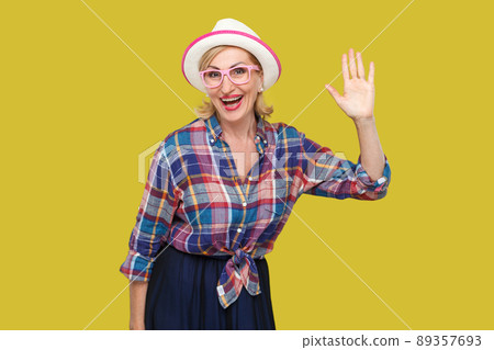 Portrait of surprised modern stylish mature woman in casual style with hat and eyeglasses standing waving her hand and looking at camera with toothy smile. studio shot isolated on yellow background. 89357693