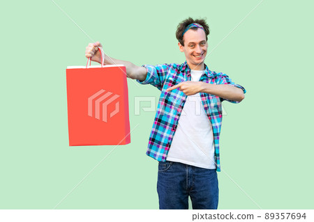 Cheerful modern young man in white t-shirt and checkered shirt standing and pointing finger to shopping bags and toothy smile, looking at camera. Studio shot, green background, isolated, indoor Cheerful modern young man in white t-shirt and checkered shirt standing and pointing finger to shopping bags and toothy smile, looking at camera. Studio shot, green background, isolated, indoor 89357694