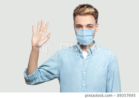 Portrait of happy excited handsome young man with surgical medical mask in blue shirt standing, looking at camera and waving his hand to greeting. indoor studio shot on grey background copy space. 89357695