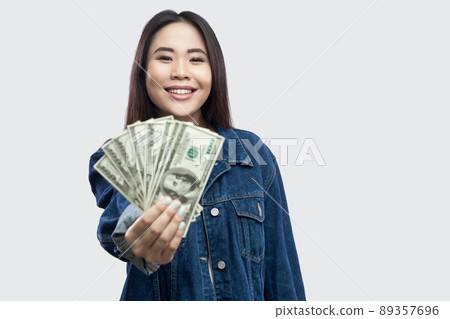 Portrait of successful attractive young asian girl in blue denim jacket standing, holding many dollars, demonstration and looking at camera with smile. Indoor, studio shot, isolated, gray background Portrait of successful attractive young asian girl in blue denim jacket standing, holding many dollars, demonstration and looking at camera with smile. Indoor, studio shot, isolated, gray background 89357696