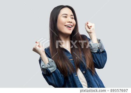 Portrait of happy surprised winner beautiful brunette asian young woman in casual blue denim jacket with makeup standing and celebrating her victory. studio shot, isolated on light grey background. 89357697