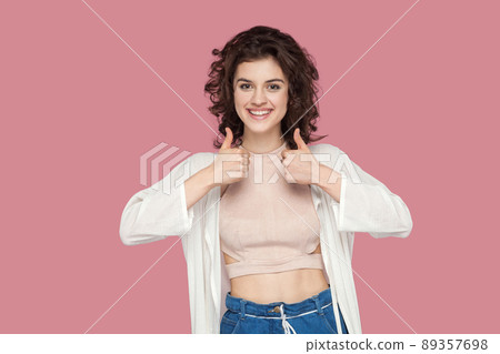 Portrait of satisfied beautiful brunette young woman with curly hairstyle in casual style standing, thumbs up and looking at camera with toothy smile. indoor studio shot isolated on pink background. 89357698