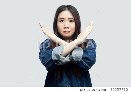 Portrait of worried beautiful brunette asian young woman in casual blue denim jacket with makeup standing and showing closed or x sign at camera. indoor studio shot, isolated on light grey background. Portrait of worried beautiful brunette asian young woman in casual blue denim jacket with makeup standing and showing closed or x sign at camera. indoor studio shot, isolated on light grey background. 89357710