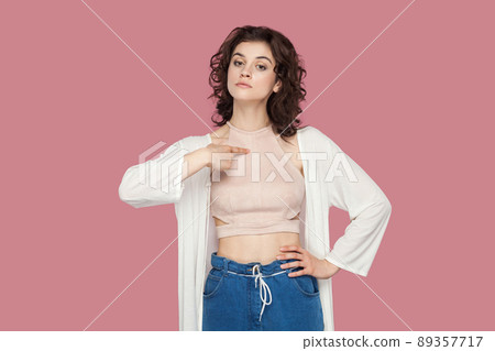 This is me. Portrait of haughty proud beautiful brunette young woman with curly hairstyle in casual style standing, pointing herself and looking at camera. studio shot isolated on pink background. 89357717