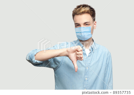 Dislike. Portrait of dissatisfied young man with surgical medical mask in light blue shirt standing, thumbs down and looking at camera with sad face . indoor studio shot on grey background copy space. Dislike. Portrait of dissatisfied young man with surgical medical mask in light blue shirt standing, thumbs down and looking at camera with sad face . indoor studio shot on grey background copy space. 89357735