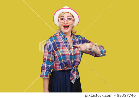 who? me? Portrait of surprised modern mature woman in casual style with hat and eyeglasses standing, looking with amazed face, pointing herself and asking. studio shot isolated on yellow background. 89357740