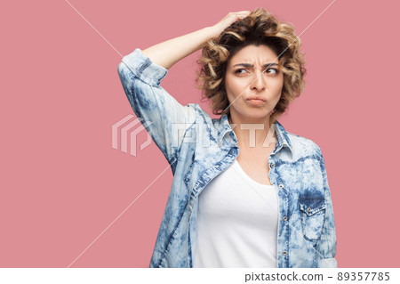 Portrait of thoughtful confused young woman with curly hairstyle in casual blue shirt standing scratching her head and thinking what to do. indoor studio shot, isolated on pink background. Portrait of thoughtful confused young woman with curly hairstyle in casual blue shirt standing scratching her head and thinking what to do. indoor studio shot, isolated on pink background. 89357785
