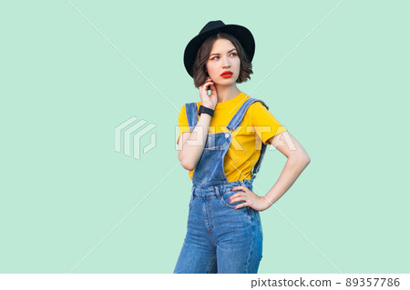 Portrait of thoughtful young hipster girl in blue denim overalls, yellow shirt, black hat standing wit hand on waist, looking away and thinking. indoor studio shot isolated on light green background. Portrait of thoughtful young hipster girl in blue denim overalls, yellow shirt, black hat standing wit hand on waist, looking away and thinking. indoor studio shot isolated on light green background. 89357786