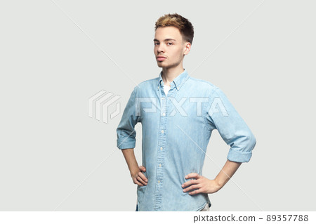 Portrait of proud satisfied handsome young man in light blue shirt standing with hands on waist and looking at camera with serious face. indoor studio shot on grey background copy space. 89357788