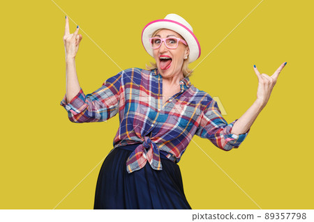 Portrait of crazy funny modern stylish mature woman in casual style with hat and eyeglasses standing, screaming and looking at camera with rock sign. indoor studio shot isolated on yellow background. Portrait of crazy funny modern stylish mature woman in casual style with hat and eyeglasses standing, screaming and looking at camera with rock sign. indoor studio shot isolated on yellow background. 89357798
