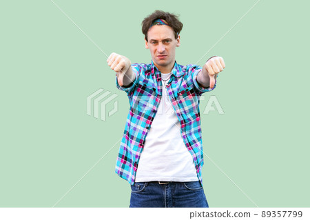 Portrait of sad young man in casual blue checkered shirt and headband standing, thumbs down and looking at camera with dislike and negative emotion. indoor studio shot, isolated on green background. Portrait of sad young man in casual blue checkered shirt and headband standing, thumbs down and looking at camera with dislike and negative emotion. indoor studio shot, isolated on green background. 89357799
